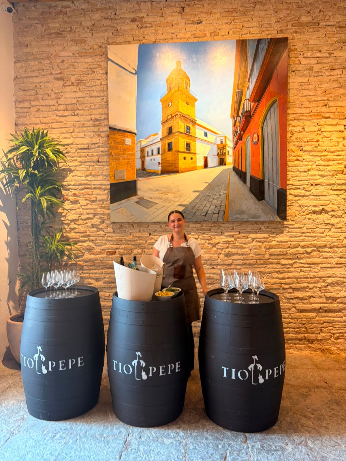 Tres barricas negras de Tío Pepe con copas de vino, atendidas por una camarera bajo el cuadro de la torre de la iglesia de Santo Domingo, sobre muro de piedra ostionera.