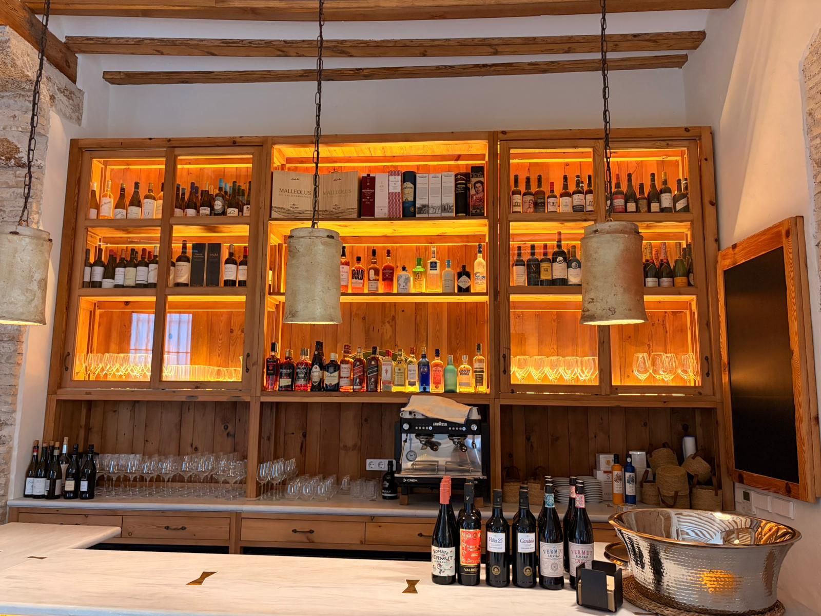 Barra de Donde Mané con vitrinas de madera retroiluminadas llenas de botellas, lámparas colgantes y vigas de madera vistas.