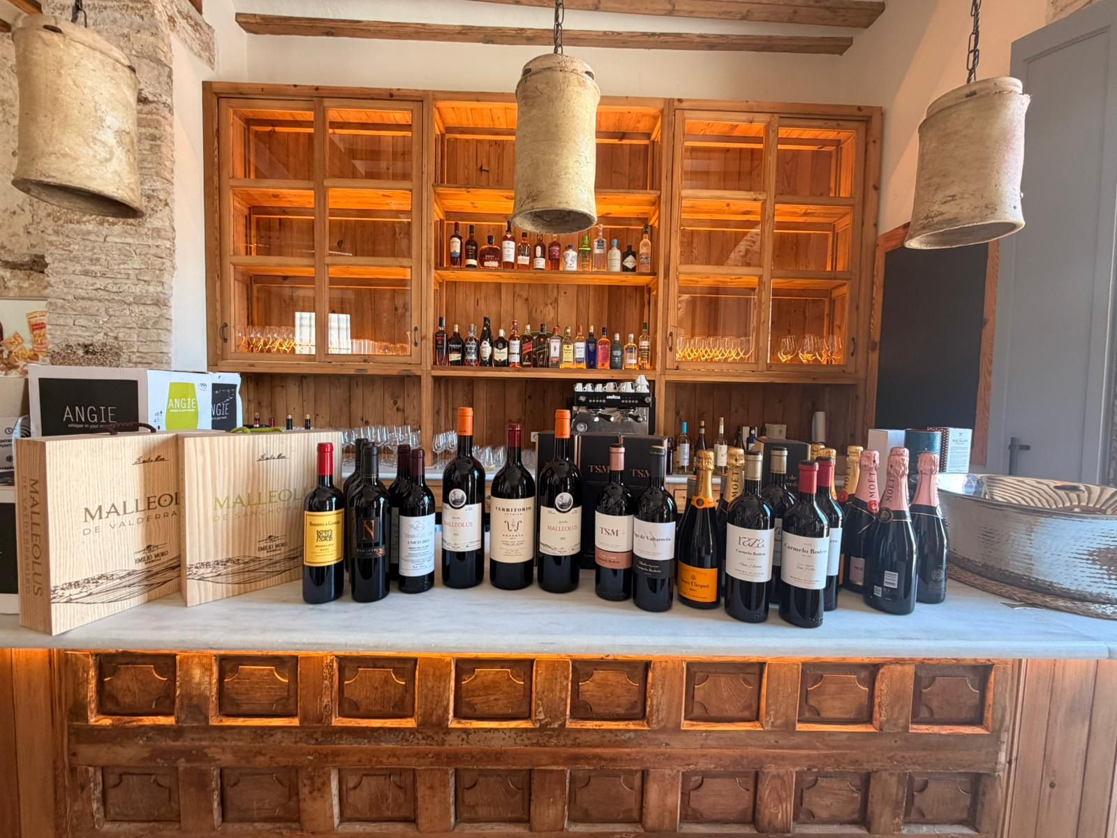 Pared de vitrinas de madera retroiluminadas con botellas de vino y una barra de mármol con botellas alineadas y cajas de madera.