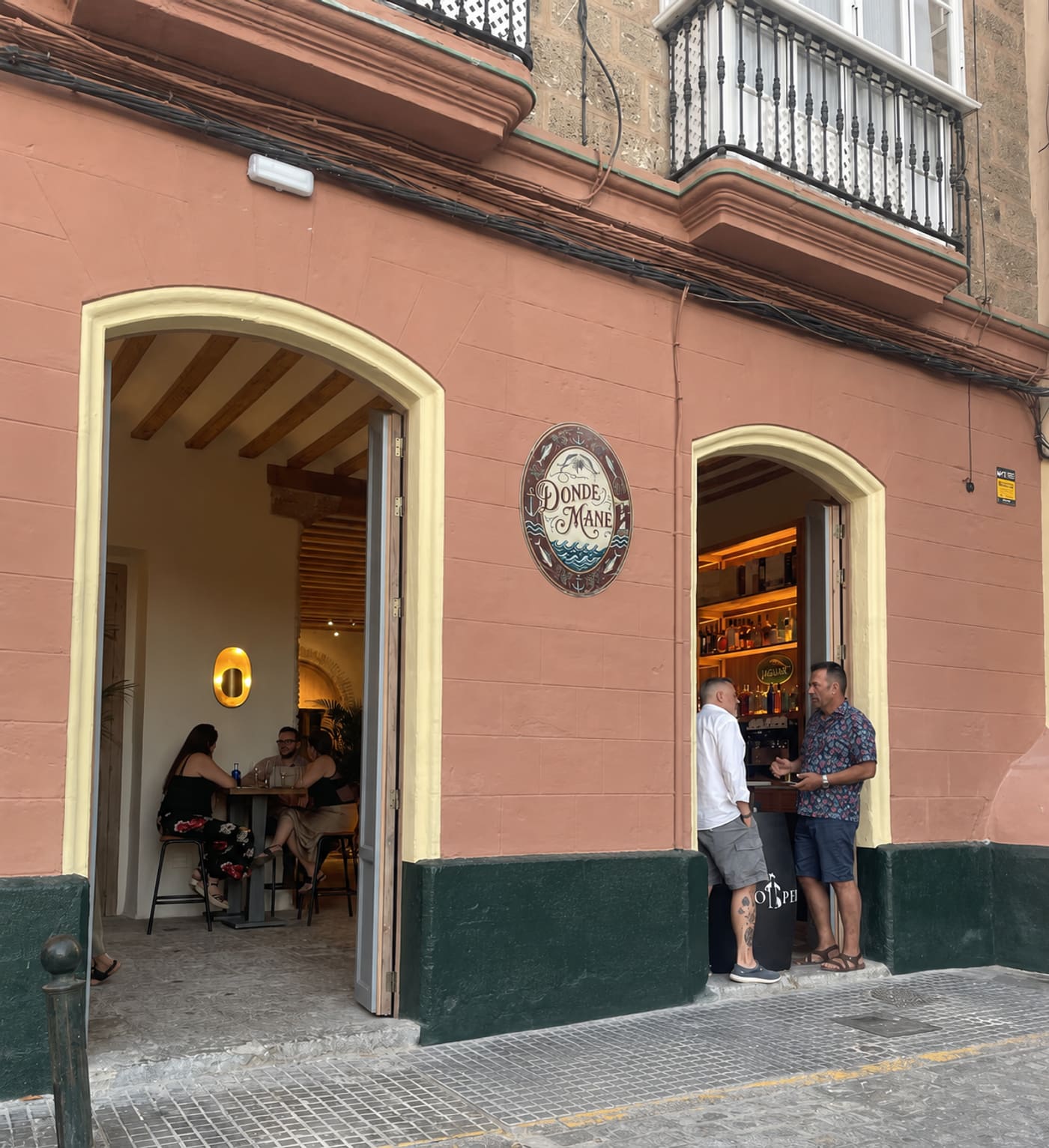 Entrada de Donde Mané: fachada salmón con zócalo verde, dos puertas en arco abiertas, el cartel ovalado entre ellas y clientes en la puerta y en el interior.