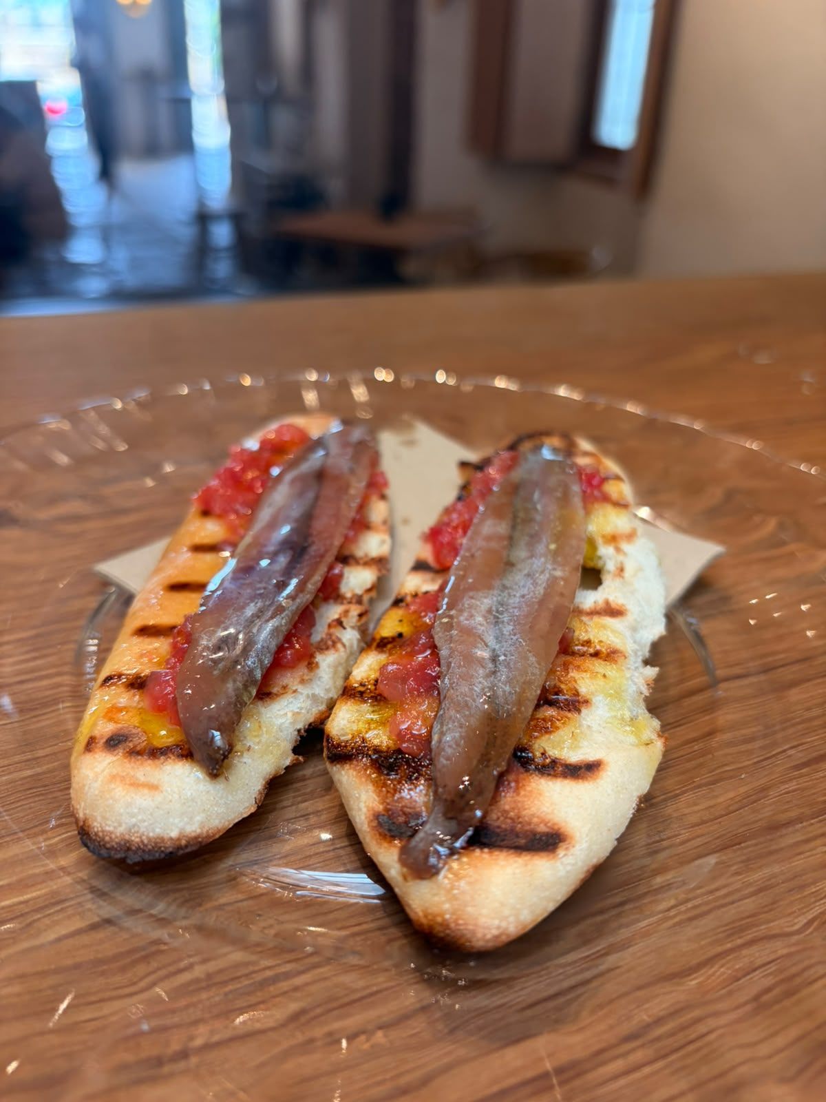 Dos montaditos de pan tostado con anchoa y tomate sobre un plato de cristal.