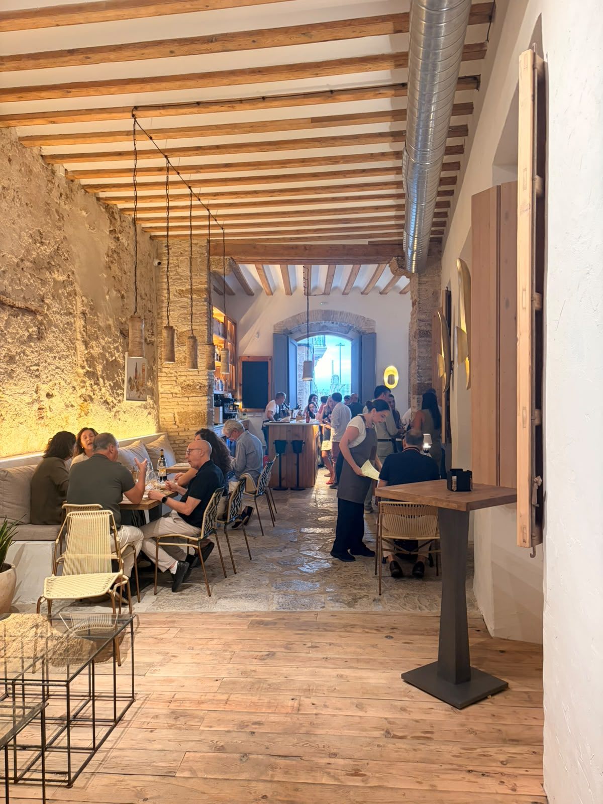 Sala alargada de Donde Mané con vigas de madera, muro de piedra, lámparas colgantes y mesas ocupadas al fondo bajo un gran arco.